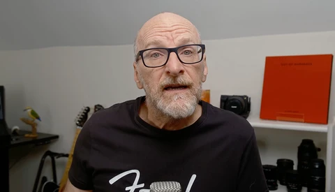 Bald man with glasses wearing a black t-shirt, speaking directly to camera in a studio setting.