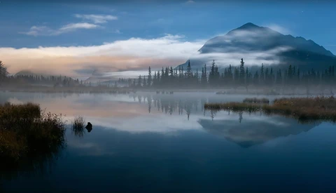 Mountain peak shrouded in mist above a forested lake at dawn, with perfect mirror reflections in still water.