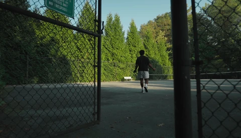 Person running through open gate onto tennis court surrounded by tall green hedges and trees.