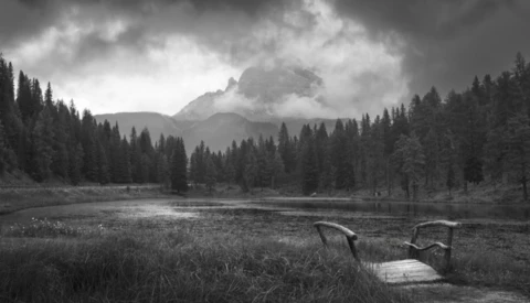 Wooden dock extending into a lake surrounded by evergreen forest with a dramatic mountain peak shrouded in clouds.
