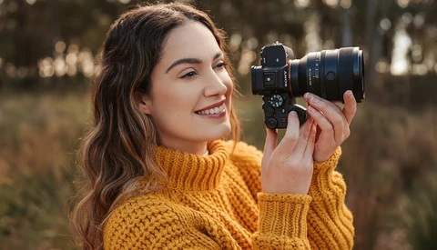 Woman in golden yellow sweater holding mirrorless camera to her eye outdoors.