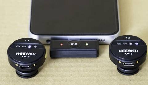 Neewer KM18 wireless microphone system with two round transmitters and central receiver connected to smartphone.