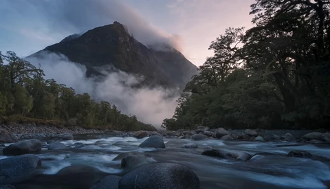 Mountain peak shrouded in mist rising above a flowing river with forested banks at twilight.