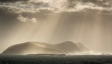 Dramatic island silhouette shrouded in atmospheric mist and volumetric light rays beneath storm clouds.