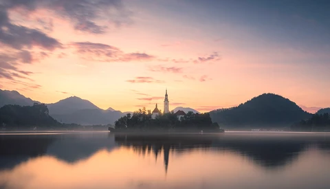 Church with spire on island in calm lake at dawn, surrounded by forested mountains.