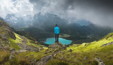 Hiker in blue jacket standing on rocky outcrop overlooking turquoise alpine lake surrounded by misty mountains.