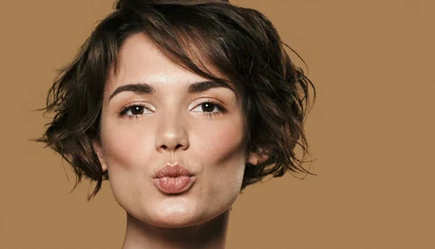Woman with short dark wavy hair making a playful kiss expression against a warm tan background.