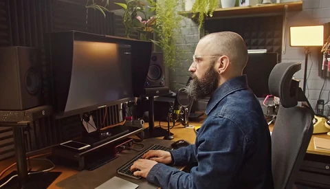 Bearded man working at a computer desk in a professional studio setup with monitor, keyboard, and audio equipment.