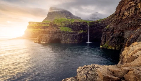 Waterfall cascading into a sea cave beneath towering coastal cliffs at golden hour.