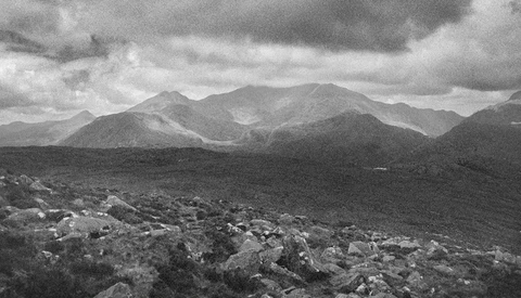 Mountain range with rocky foreground under dramatic cloudy sky in black and white.