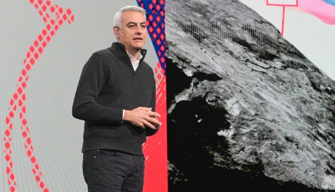 Speaker gesturing while presenting on stage with abstract geometric graphics and a black and white mountain photograph displayed on large screens behind him.