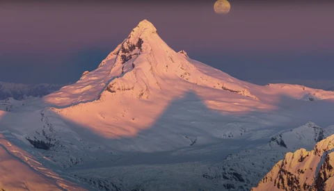 Snow-covered conical volcano peak illuminated by golden sunrise light, with the moon visible above and surrounding snowy terrain below.
