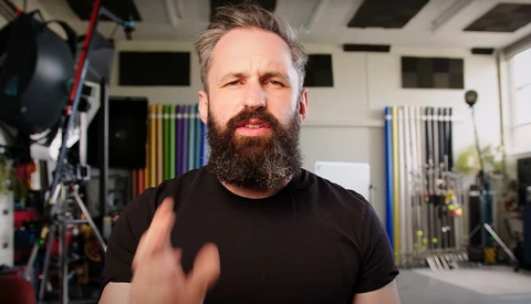 Bearded man in black t-shirt gesturing while speaking in a studio with professional lighting equipment visible.
