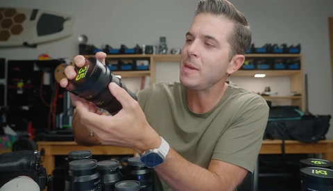 Man in tan shirt examining a black camera lens in a workshop setting.