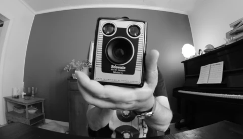 Photographer holds a vintage square-format camera with dual lenses toward the camera in a black and white self-portrait.