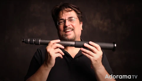 Man wearing glasses holding a telephoto lens toward the camera against a dark background.