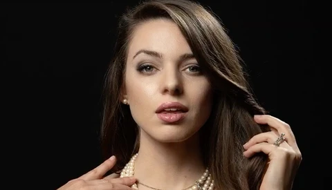 Woman with long dark hair wearing pearl necklace and ring, posed against black background.