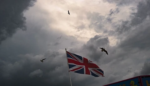 Union Jack flag rippling against dramatic cloudy sky with birds in flight.