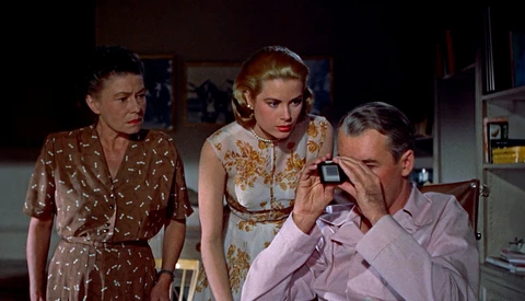 Three people in a 1950s interior setting, with a man holding a camera while two women observe.
