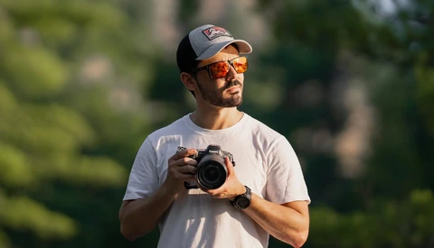 Male photographer holding a DSLR camera outdoors in natural daylight, wearing a cap and orange-tinted sunglasses.
