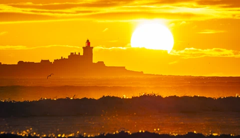 Silhouetted lighthouse and coastal town against brilliant golden sunset with large sun on horizon.