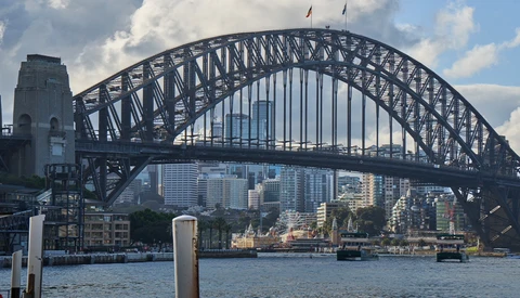Steel arch bridge spanning a harbor with modern city skyline and waterfront buildings visible beneath it.