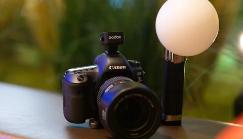Canon DSLR camera with Godox flash and diffuser dome mounted on top, set against blurred outdoor background.