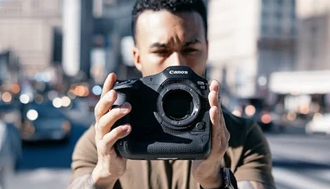 Photographer holding Canon EOS R5 camera body in urban setting with blurred cityscape background.