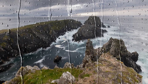 Rocky coastline with turquoise water viewed through rain-covered glass.