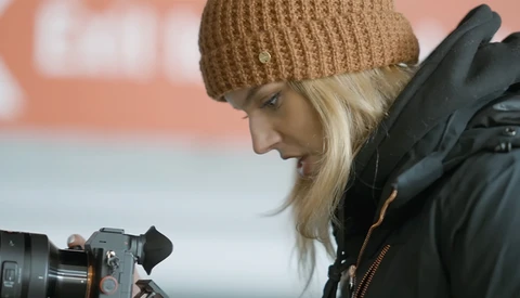 Female videographer with professional camera wearing tan beanie and black jacket.