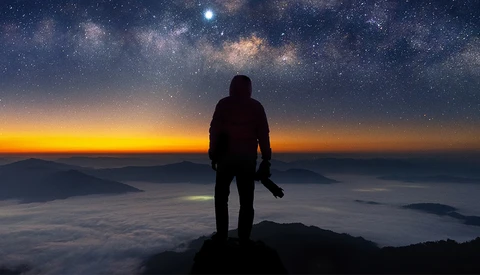Silhouetted figure standing on a mountain peak overlooking clouds at sunrise with the Milky Way visible above.