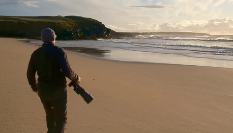 Person in dark jacket walking across sandy beach toward rolling waves with green headland in background.