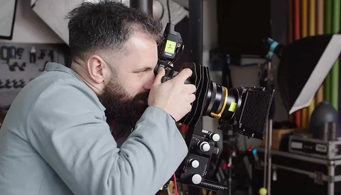 Bearded photographer looking through large format camera viewfinder in studio setting.