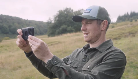 Man in cap holding smartphone to capture landscape photo in rolling hills.