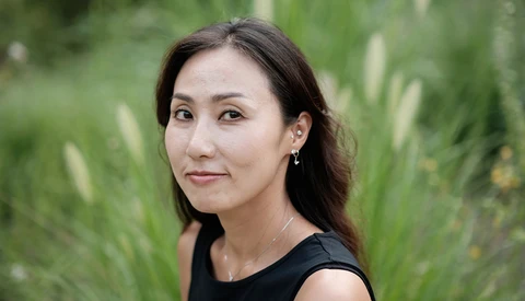 Woman with long dark hair wearing black sleeveless top, posed outdoors against soft green foliage background.
