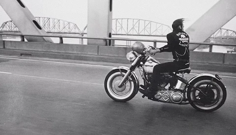 Motorcyclist riding a cruiser on a bridge with architectural steel framework visible in the background.