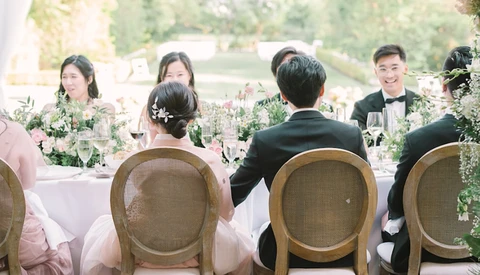 Wedding reception table with bride and groom seated at head table with wedding party members, surrounded by floral centerpieces and greenery.