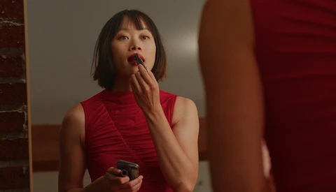 Woman in red sleeveless turtleneck applying lipstick while looking in mirror.