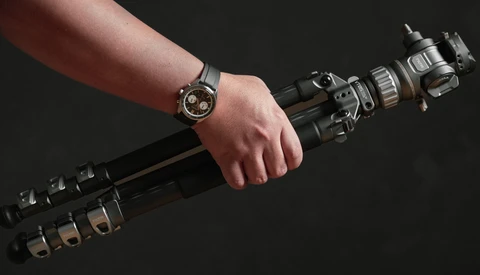 Photographer's hand gripping a telephoto lens with a watch visible on the wrist against a dark background.
