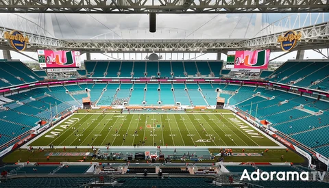 Large stadium with turquoise seats and green field viewed from upper seating level under steel roof structure.