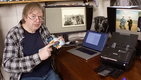 Photographer holding a light meter while seated at a desk with a laptop and scanner.
