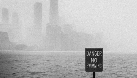 A "Danger No Swimming" sign stands in the foreground beside foggy water with a city skyline barely visible in heavy mist.