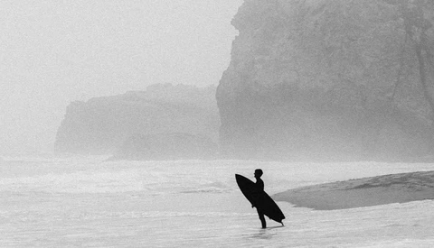 Surfer in wetsuit standing in shallow water holding surfboard, facing misty cliffs.