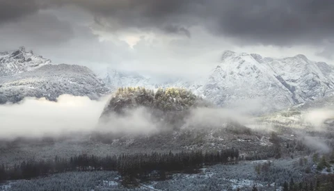 Snow-capped mountains frame a forested valley shrouded in dense fog and mist.