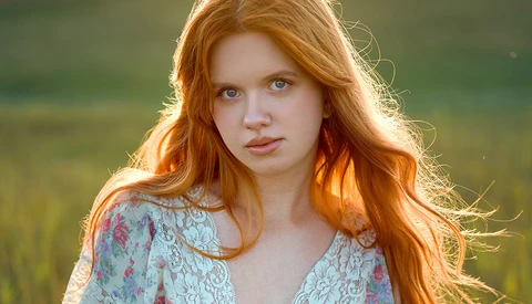 Portrait of a young woman with long red hair wearing a floral lace top, backlit by golden hour sunlight.