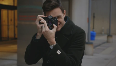 Young man in black jacket holding a rangefinder camera to his face, smiling while shooting in an urban corridor.