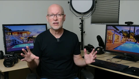 Man in black shirt gesturing while seated at a studio desk with photography equipment and monitors displaying architectural images.