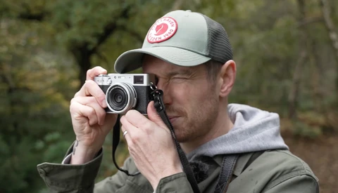Photographer holding a rangefinder camera up to his eye while wearing a cap and olive jacket.