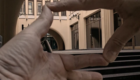 Hands framing a courtyard view through a window opening, with classical architecture visible beyond.