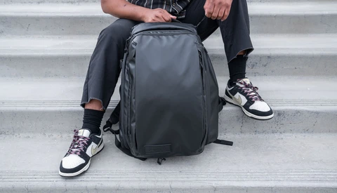 Person sitting on concrete steps holding a dark gray backpack with organizational compartments.